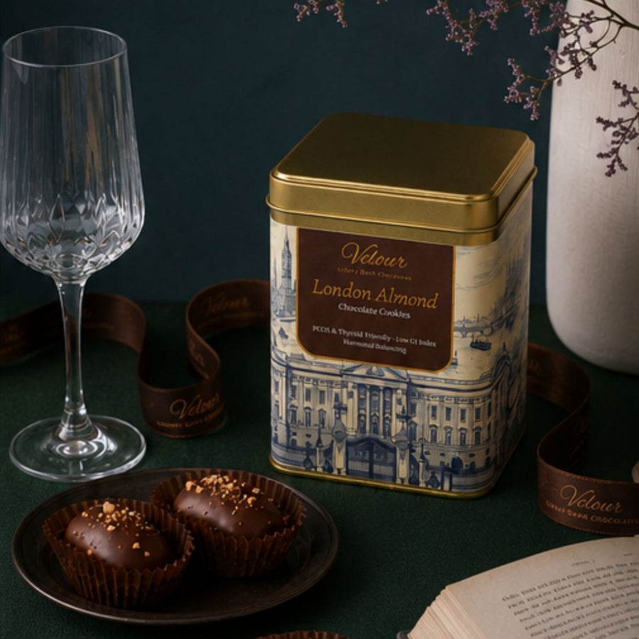 Velour London Almond chocolate cookie tin with a glass of wine and cookies on a dark surface.