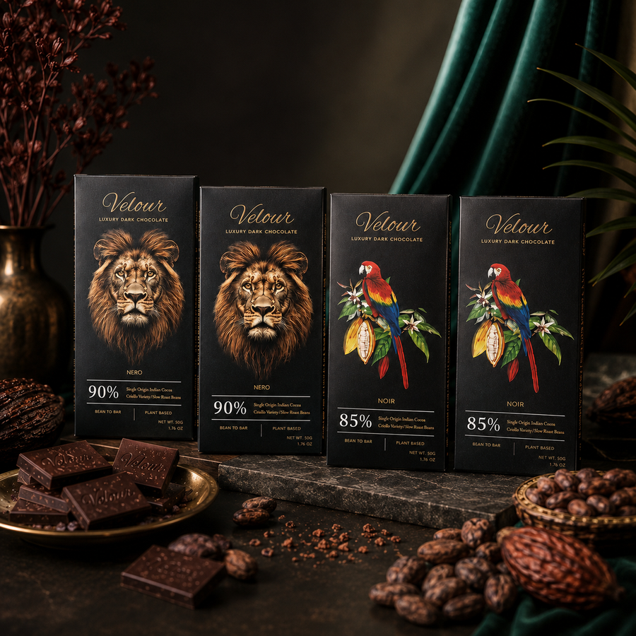 Four black chocolate bars from the brand Vélour are displayed on a dark stone surface with chocolate pieces scattered around. The packaging features artistic illustrations of a lion, two parrots, and a toucan, with dark chocolate pieces and cacao beans used as styling props against a dark, moody background with green drapery.